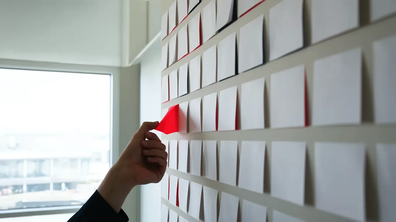 Hand setzt einen roten Notizzettel in ein Raster weißer Zettel an einer Wand